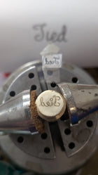 A close-up of a metal engraving vise holding a round brass piece engraved with “herz.” Above is a template, and in the background, a blurry sheet reads “Tied”—the perfect Tied Charm by Jessica de Lotz Jewellery for friendship jewelry or wax seals.