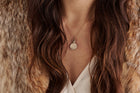 A woman with long, wavy brown hair wears a fur coat over a white textured top. At her collarbone rests the Jessica de Lotz Jewellery House of the Dragon & JdL Targaryen Wax Seal Charm, its engraved pendant elegant and round.