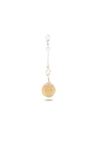 A silver keychain with a short chain and clasp holds Jessica de Lotz Jewellery’s Midnight Margarita Lime Charm—a round, gold-tone medallion adorned with a stylized sunburst design. Reminiscent of modern-day chatelaine accessories, this piece channels the spirit of Practical Magic charms as the medallion dangles from the end of the chain, casting a delicate shadow on a white background.