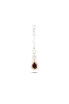 A silver earring with a delicate chain and hook, featuring a teardrop-shaped deep red gemstone set in a silver bezel—this elegant piece, the “My Blood, Your Blood, Our Blood Charm” by Jessica de Lotz Jewellery, draws inspiration from sisterhood jewelry and evokes the mystical bonds reminiscent of Practical Magic charms. The background is plain white with a faint shadow beneath the earring.
