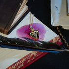 An Amas Veritas Spell Charm by Jessica de Lotz Jewellery, a gold heart-shaped locket on a chain, rests atop a lined notebook alongside a purple flower, black feather, an old book, and envelopes on a dark surface. Reminiscent of the sisterhood charms from Practical Magic 2, the scene evokes a vintage and sentimental atmosphere—like uncovering a modern chatelaine brimming with secrets and memories.