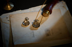 Two vintage gold pocket watches, one on a chain, rest atop worn, stacked envelopes. The softly lit scene features the Jessica de Lotz Jewellery Verbena Botanical Apothecary Bottle Charm and an old stamp nearby, evoking a nostalgic, historical atmosphere. Shadows and faded ink lines enhance the antique feel.