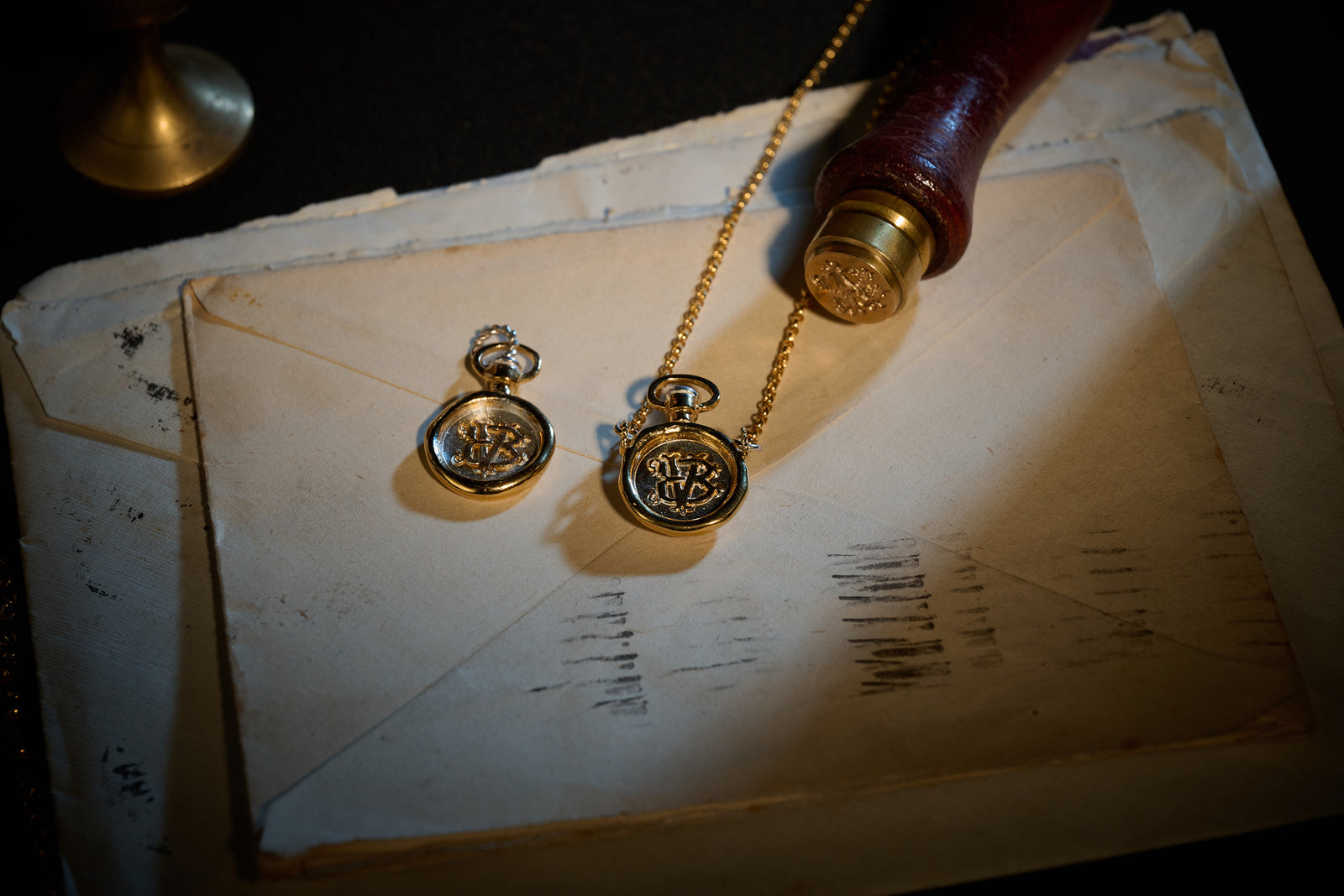 Two vintage gold pocket watches, one on a chain, rest atop worn, stacked envelopes. The softly lit scene features the Jessica de Lotz Jewellery Verbena Botanical Apothecary Bottle Charm and an old stamp nearby, evoking a nostalgic, historical atmosphere. Shadows and faded ink lines enhance the antique feel.