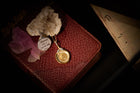 A Midnight Margarita Charm necklace by Jessica de Lotz Jewellery, featuring gold and silver detailing, rests on a crystal atop an old textured book. Nearby, pink rose petals and a folded, yellowed piece of handwritten paper add to the scene, all set against a dark background with dramatic lighting that evokes the enchantment of Practical Magic-inspired charms.