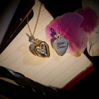 Two heart-shaped lockets from Jessica de Lotz Jewellery rest on an open notebook, channeling a modern-day chatelaine: one gold locket is adorned with a delicate leaf motif, while the other—the Amas Veritas Spell Charm—gleams in silver and bears its namesake engraving. Both pendants hang from graceful chains like tokens of sisterhood passed through generations. Scattered pink flower petals and vintage letters nearby bask in dramatic, magical light reminiscent of the world seen in Practical Magic 2.