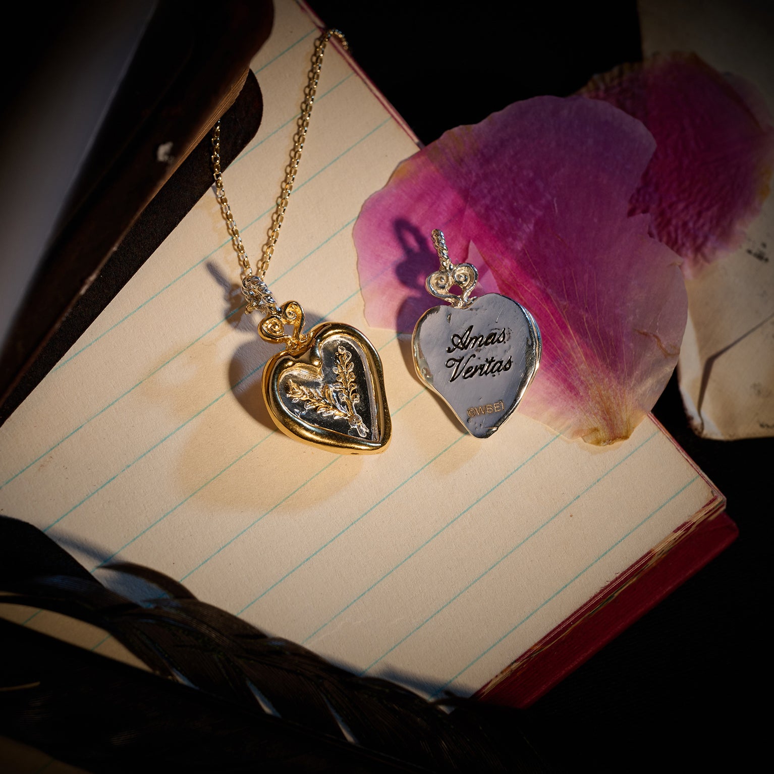 Two heart-shaped lockets from Jessica de Lotz Jewellery rest on an open notebook, channeling a modern-day chatelaine: one gold locket is adorned with a delicate leaf motif, while the other—the Amas Veritas Spell Charm—gleams in silver and bears its namesake engraving. Both pendants hang from graceful chains like tokens of sisterhood passed through generations. Scattered pink flower petals and vintage letters nearby bask in dramatic, magical light reminiscent of the world seen in Practical Magic 2.