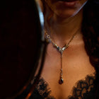 A woman gazes into a mirror, her reflection softly blurred as she wears the Jessica de Lotz Jewellery “My Blood, Your Blood, Our Blood Charm” gold necklace with its striking red pendant. Dim lighting accentuates her lower face, neck, and a lace-trimmed black top, while the mirror subtly captures both her features and the enchanting jewelry.