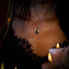 A woman wearing a delicate gold necklace adorned with the “My Blood, Your Blood, Our Blood Charm” by Jessica de Lotz Jewellery—its striking blue teardrop pendant partially visible against a low-cut black lace top. Soft candlelight bathes her skin and the lace in a warm, intimate glow.