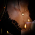 A woman wearing a lace-trimmed black garment and a Jessica de Lotz Jewellery Verbena Botanical Apothecary Bottle Charm necklace is seen in soft candlelight. Several lit white candles are in the foreground, creating a warm, intimate ambiance. Her face is not visible in the image.