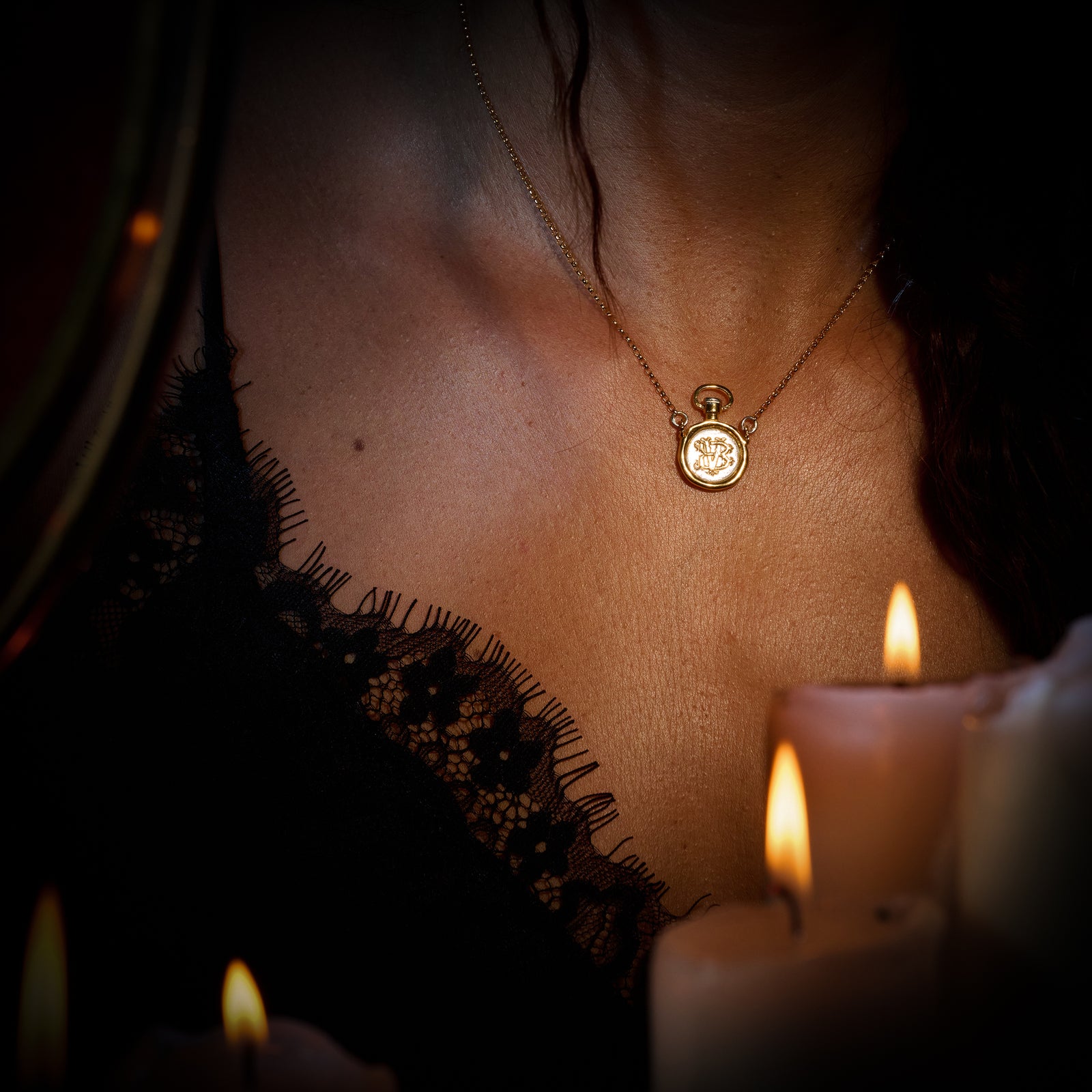 A woman wearing a lace-trimmed black garment and a Jessica de Lotz Jewellery Verbena Botanical Apothecary Bottle Charm necklace is seen in soft candlelight. Several lit white candles are in the foreground, creating a warm, intimate ambiance. Her face is not visible in the image.