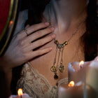 A woman wearing a lace-trimmed top touches her chest, showcasing a ring and the Jessica de Lotz Jewellery Midnight Margarita Charm—a silver chatelaine hand necklace adorned with whimsical accents. Lit candles in the foreground cast a warm, enchanting glow.