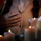 A woman dressed in lace gently touches her chest, showcasing the Jessica de Lotz Jewellery Chatelaine Hand Necklace with ‘Our Blood’ Drop Charm. Her manicured nails and a ring add to her elegant look, while several lit candles in the foreground bathe the scene—reminiscent of Practical Magic 2—in a warm, intimate glow. Wax seal charms nearby enhance the soft and enchanting atmosphere.
