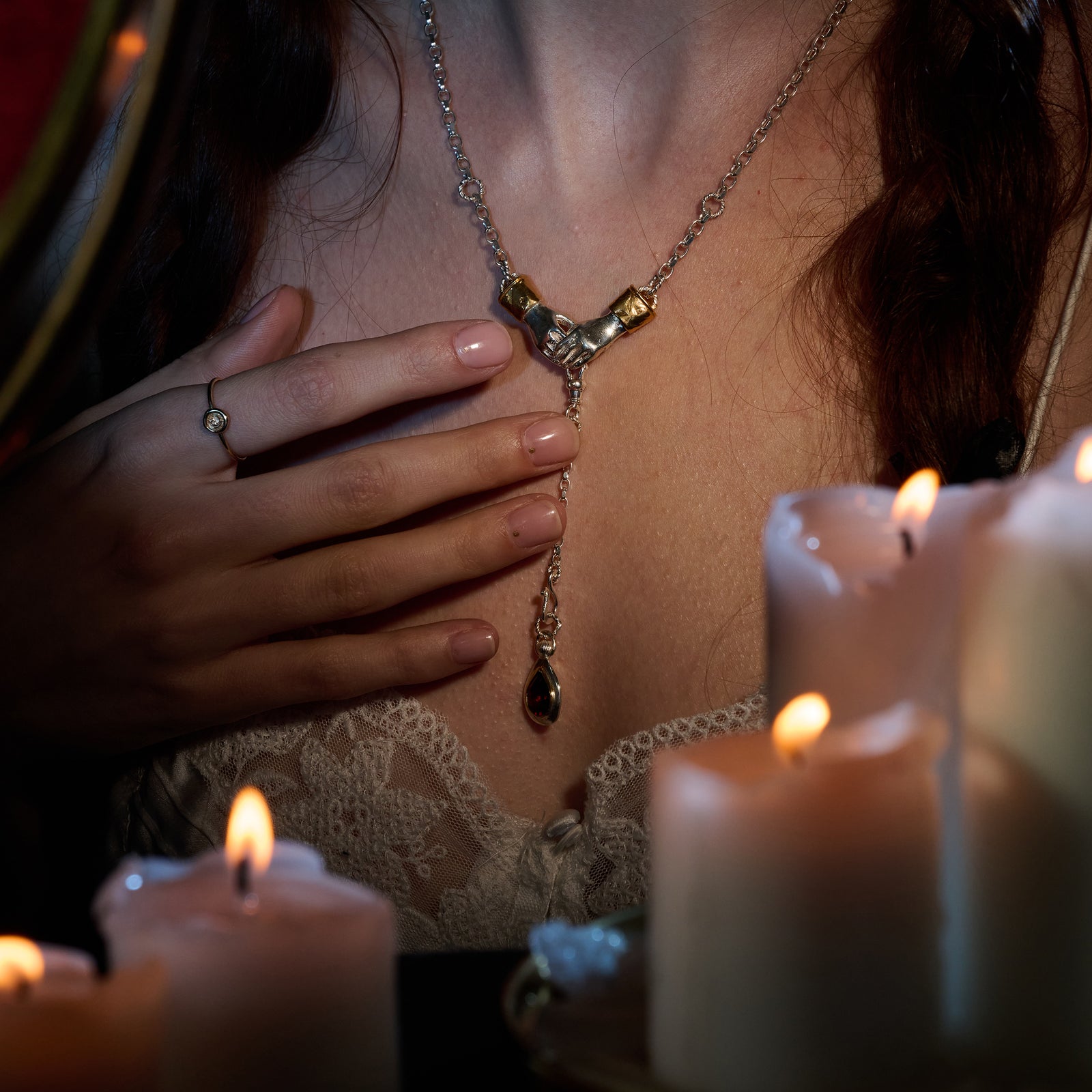 A woman dressed in lace gently touches her chest, showcasing the Jessica de Lotz Jewellery Chatelaine Hand Necklace with ‘Our Blood’ Drop Charm. Her manicured nails and a ring add to her elegant look, while several lit candles in the foreground bathe the scene—reminiscent of Practical Magic 2—in a warm, intimate glow. Wax seal charms nearby enhance the soft and enchanting atmosphere.