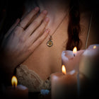 A woman with braided hair wears a lace-trimmed top, holding the Jessica de Lotz Jewellery Verbena Botanical Apothecary Bottle Charm close to her chest. She has a simple ring on her finger—a delicate piece symbolizing sisterhood. Several lit, partially melted candles glow in the foreground, casting an enchanting light reminiscent of Practical Magic charms.