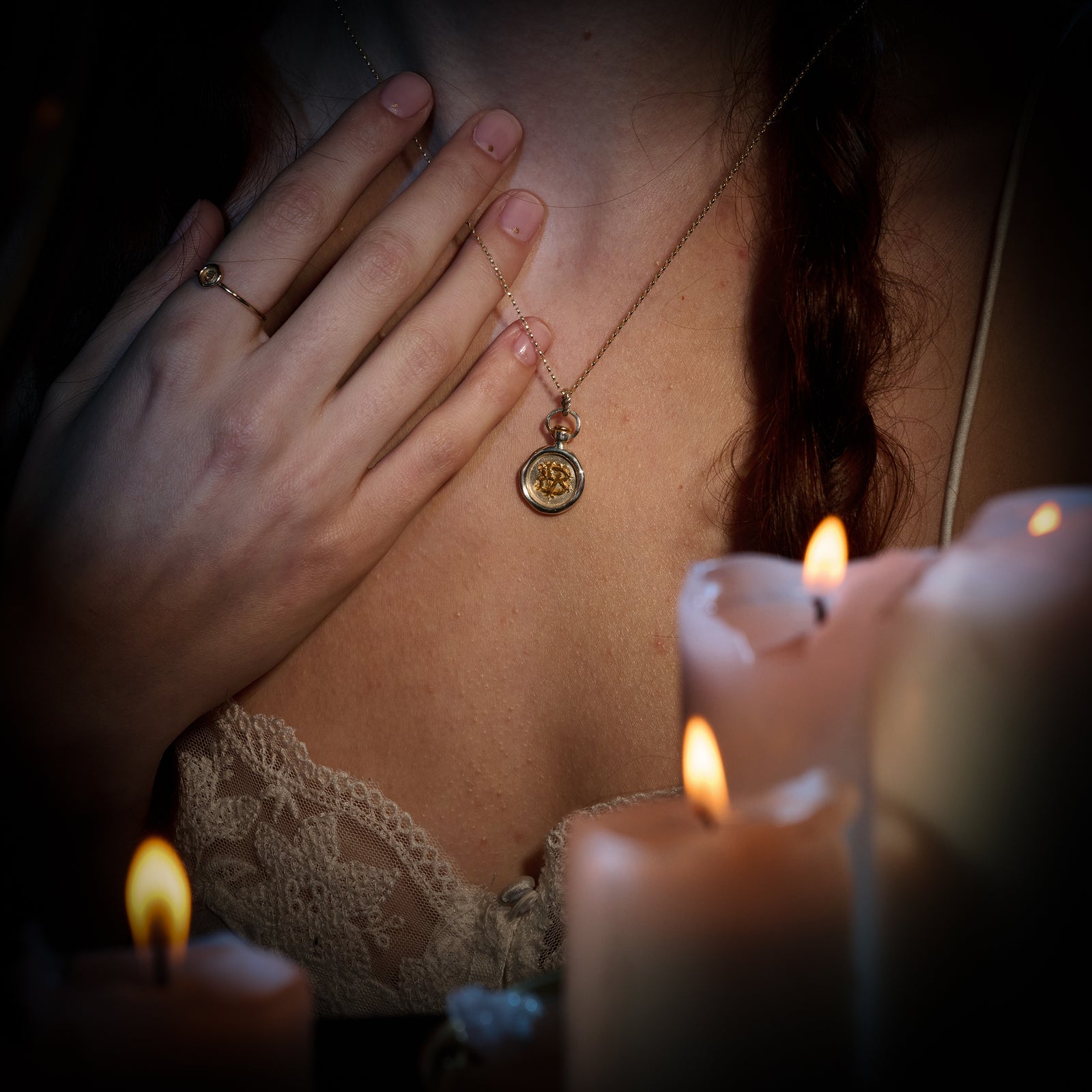 A woman with braided hair wears a lace-trimmed top, holding the Jessica de Lotz Jewellery Verbena Botanical Apothecary Bottle Charm close to her chest. She has a simple ring on her finger—a delicate piece symbolizing sisterhood. Several lit, partially melted candles glow in the foreground, casting an enchanting light reminiscent of Practical Magic charms.