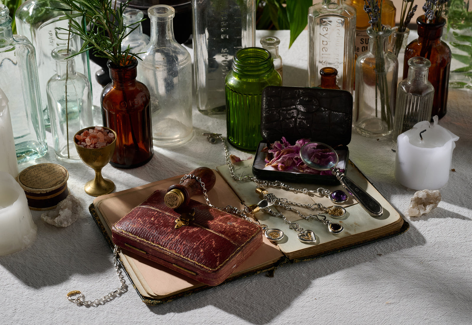 An arrangement featuring vintage glass bottles, a candle, a red leather journal, a gold goblet filled with pink salt, a silver magnifying glass, sisterhood jewelry by Jessica de Lotz Jewellery, the Verbena Botanical Apothecary Bottle Charm by Jessica de Lotz Jewellery scattered among botanical wax-seal charms, dried flowers, and crystals rests on a sunlit white tablecloth.