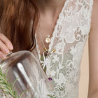 A person in a white, floral lace top holds a glass cloche over a sprig of rosemary. They have long, reddish-brown hair and wear layered silver necklaces, including a delicate botanical piece—possibly the Verbena Botanical Apothecary Bottle Charm Necklace ~ Limited Edition from Jessica de Lotz Jewellery—with one chain featuring a wax-seal charm and another adorned with a purple bead.