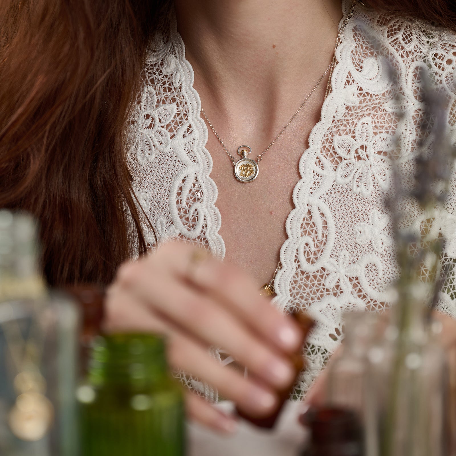 A woman with long brown hair wears an intricately scalloped white lace top and the limited edition Verbena Botanical Apothecary Bottle Charm Necklace by Jessica de Lotz Jewellery. As her hand reaches for a small amber glass bottle amid other glass jars on the table, attention is drawn to her delicate botanical jewelry—the distinctive necklace inspired by apothecary bottles—and her detailed lace clothing.