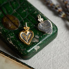 Two heart-shaped Amas Veritas Spell Charms by Jessica de Lotz Jewellery rest atop a green vintage box: one in gold featuring a delicate leaf motif, the other in silver engraved with “Amor Veritas.” Reminiscent of modern chatelaine, these sisterhood charms each have small loop bails and are displayed on fabric beside an old letter and a sprig of lavender—a scene evocative of the enchanting world of Practical Magic 2.