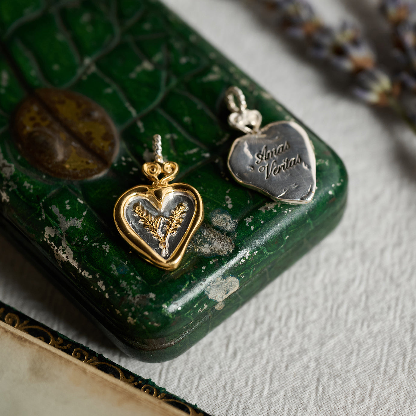 Two heart-shaped Amas Veritas Spell Charms by Jessica de Lotz Jewellery rest atop a green vintage box: one in gold featuring a delicate leaf motif, the other in silver engraved with “Amor Veritas.” Reminiscent of modern chatelaine, these sisterhood charms each have small loop bails and are displayed on fabric beside an old letter and a sprig of lavender—a scene evocative of the enchanting world of Practical Magic 2.