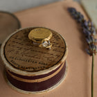 A small round box with a brown ribbon edge sits on a peach surface. On top rests a gold locket, a tiny silver rose pin, and the Verbena Botanical Apothecary Bottle Charm by Jessica de Lotz Jewellery—a delicate piece reminiscent of nature-inspired keepsakes. Handwritten text and an address are visible on the box lid. A sprig of lavender lies to the right, evoking the spirit of sisterhood jewelry and vintage treasures.