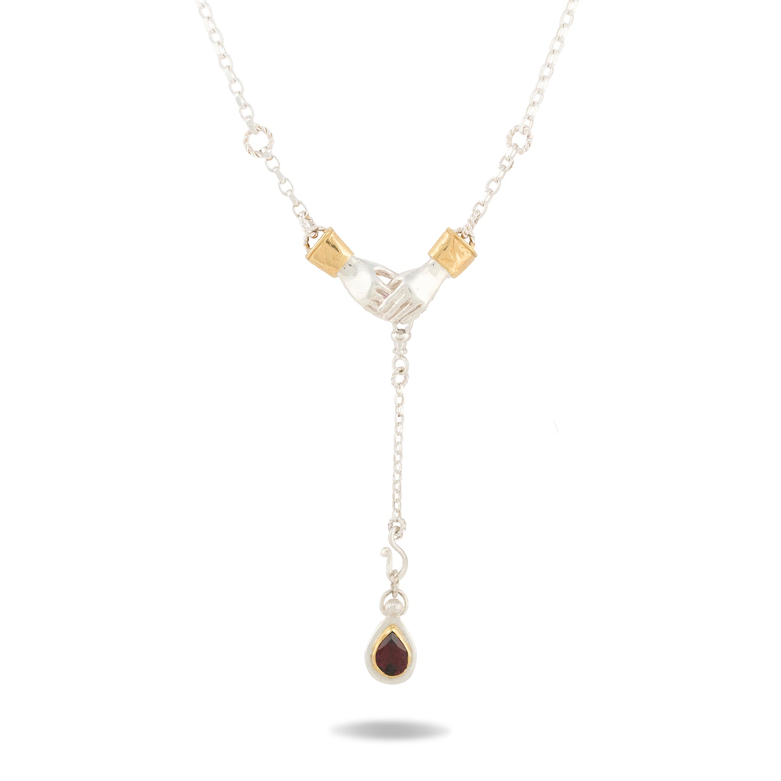 The Chatelaine Hand Necklace with ‘Our Blood’ Drop Charm by Jessica de Lotz Jewellery features two silver clasped hands with gold cuffs, joined by a delicate chain. Suspended below is a vertical drop ending in a teardrop-shaped, dark red gemstone framed in silver—evoking the mystical aesthetic of Practical Magic. Set against a plain white background, the pendant casts a subtle shadow.