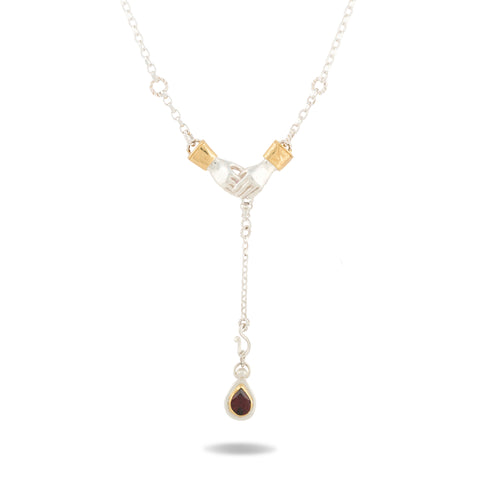 The Chatelaine Hand Necklace with ‘Our Blood’ Drop Charm by Jessica de Lotz Jewellery is a limited edition piece featuring two silver hands clasped together, each adorned with gold cuffs. A delicate chain with small, evenly spaced links gracefully suspends a teardrop-shaped dark red garnet charm set in silver. This elegant necklace symbolizes lasting bonds and sisterhood.