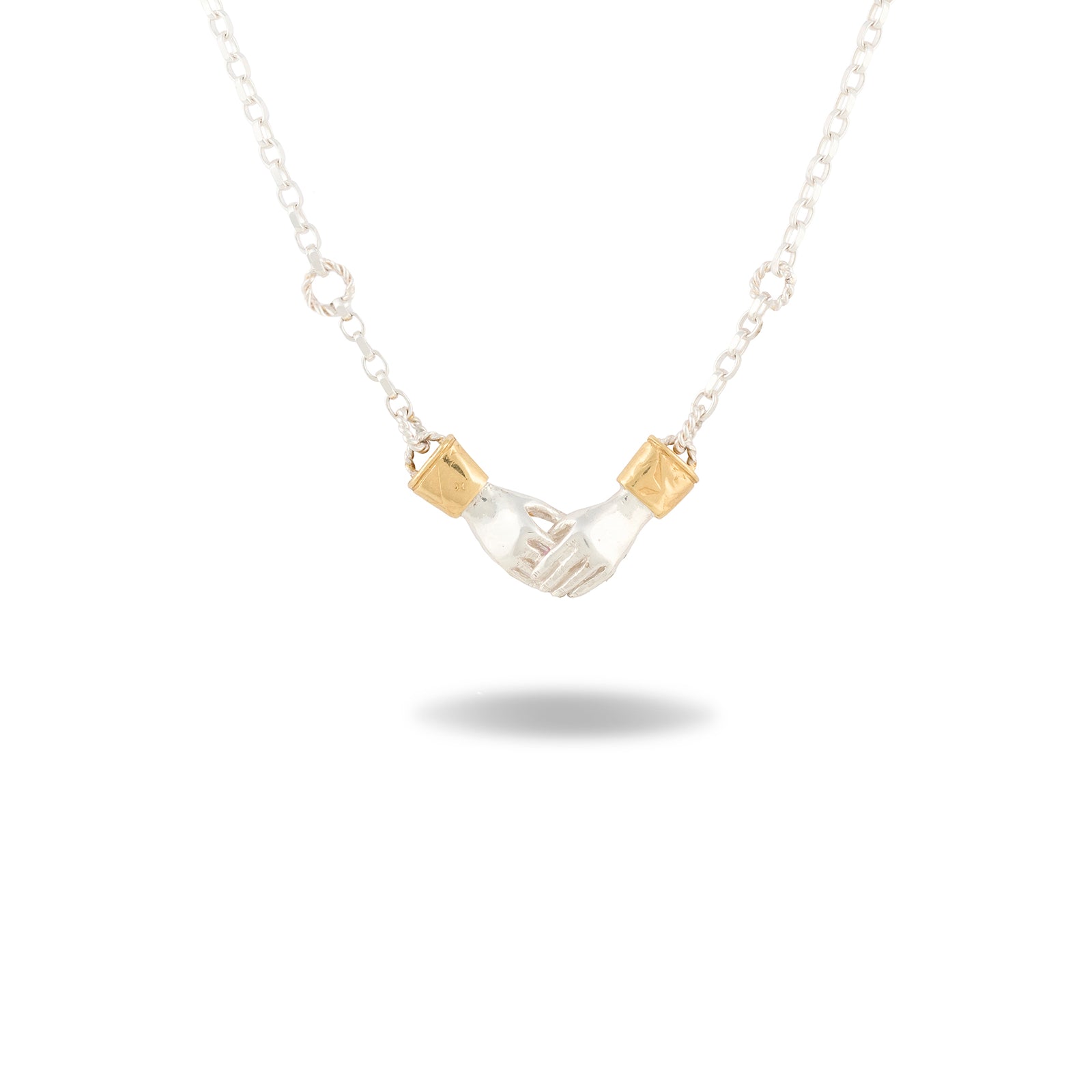 The Chatelaine Hand Necklace by Jessica de Lotz Jewellery features a pendant of two clasped silver hands, each cuffed with a petite gold band. Suspended from a delicate silver chain and set against a plain white background with a soft shadow beneath, this piece evokes the timeless essence of Practical Magic jewelry.