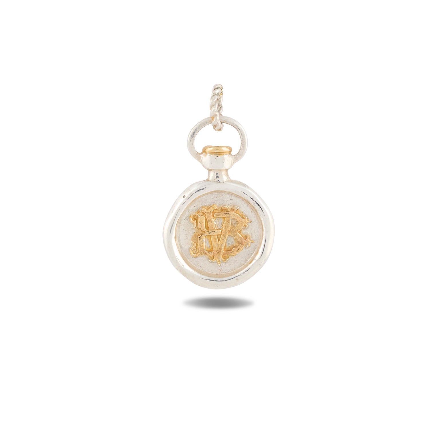 A small, round silver Verbenca Botanical Charm Bottle Top by Jessica de Lotz Jewellery features a twisted loop at the top. Reminiscent of Practical Magic charms, its center displays an ornate gold monogram with the letters V and B intertwined. The pendant casts a faint shadow on the white background.