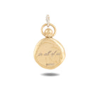 A round, gold pendant from Jessica de Lotz Jewellery called “There’s A Little Witch In All Of Us Charm,” reminiscent of a wax seal with a small loop at the top and engraved with the phrase “in all of us” followed by OWBEI on its front. The charm casts a slight shadow on the white background.
