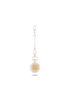 A Verbena Botanical Apothecary Bottle Charm by Jessica de Lotz Jewellery, featuring a gold-tone round pendant with an ornate relief design, is suspended from a short decorative silver chain with a clasp. Reminiscent of sisterhood jewelry and imbued with the enchanting essence of Practical Magic-inspired charms, this piece is beautifully showcased against a white background, its delicate shadow cast beneath it.