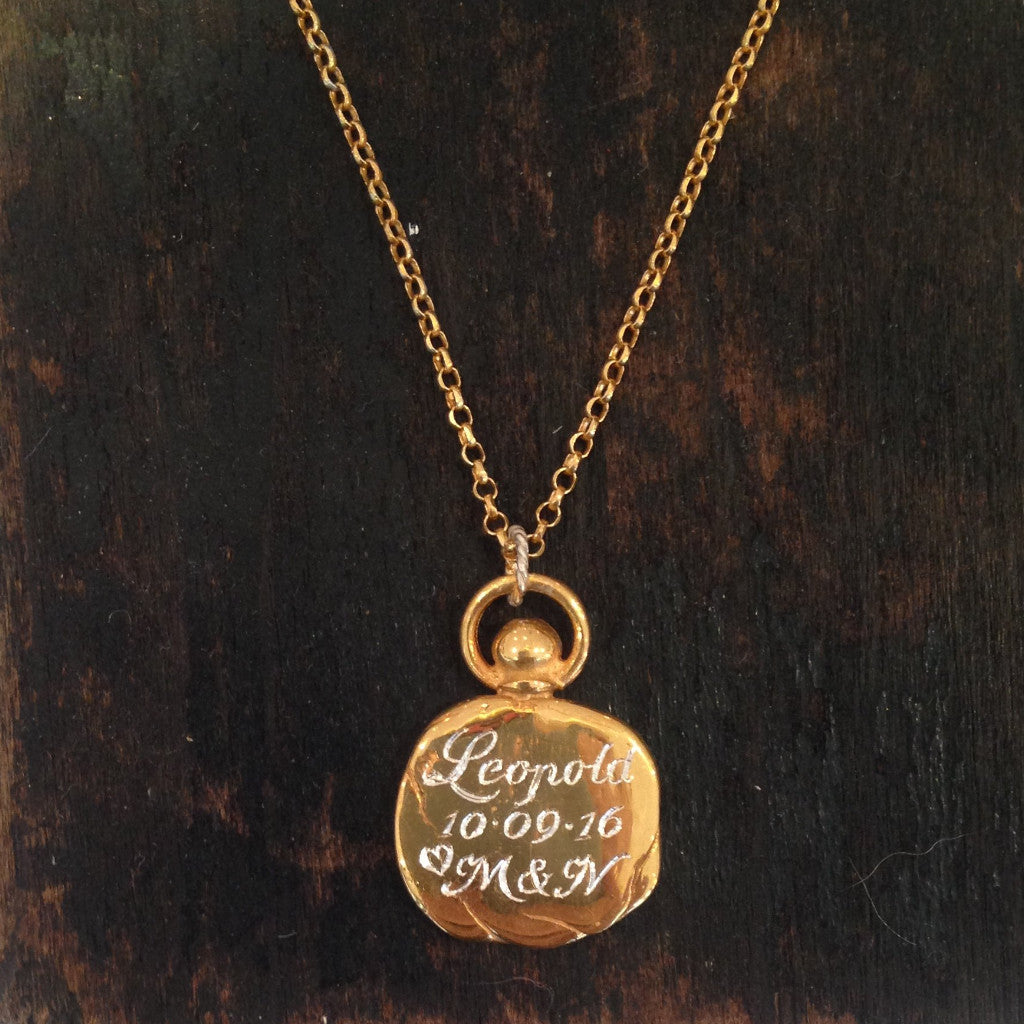 A Hand or Paw Imprint Charm by Jessica de Lotz Jewellery rests on a dark wooden background. This personalised piece features engraved details in cursive, with its delicate chain neatly centered in the image.