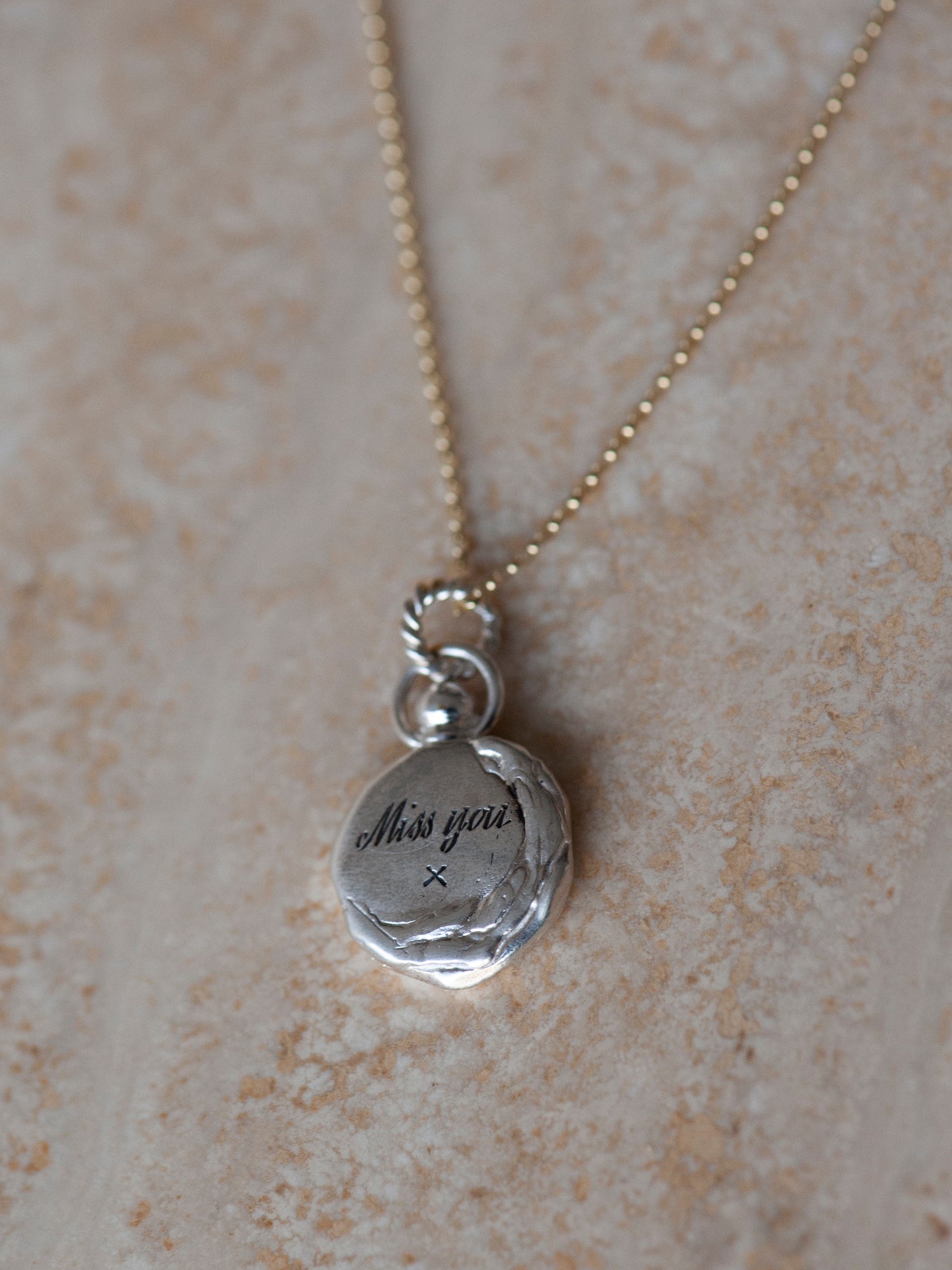 A close-up of the Jessica de Lotz Jewellery Hand or Paw Imprint Charm, a round silver pendant styled like a wax seal on a gold chain, engraved with "Miss you" and an x, displayed on a textured beige surface.