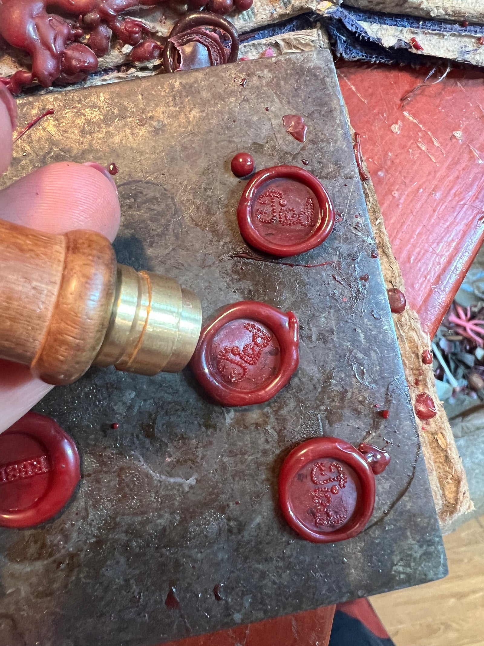 A hand presses the Jessica de Lotz Jewellery Tied Charm—a brass and wood seal stamp—into red wax on a metal plate, creating unique round seals. Finished charms and wax drips decorate the worn metal surface and rich red wooden table.