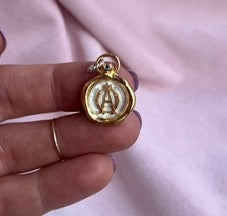 A Jessica de Lotz Jewellery Design Your Own: 2D Art Charm held between two fingers, featuring a cream-colored wax-seal style charm with a gold rim, an embossed capital letter A, and a small jump ring at the top.