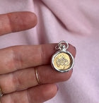 A hand holds a Design Your Own: Engraved Symbolic Charm by Jessica de Lotz Jewellery, featuring a gold-toned floral motif encased in a silver border with a jump ring.