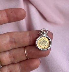 A hand holds a Design Your Own: Engraved Symbolic Charm by Jessica de Lotz Jewellery, featuring a gold-toned floral motif encased in a silver border with a jump ring.