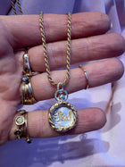 A hand adorned with gold rings holds Jessica de Lotz Jewellery's Tied Charm—a gold chain necklace with a round, wax seal-style pendant engraved with "TIED" on white—resting against pale pink fabric.