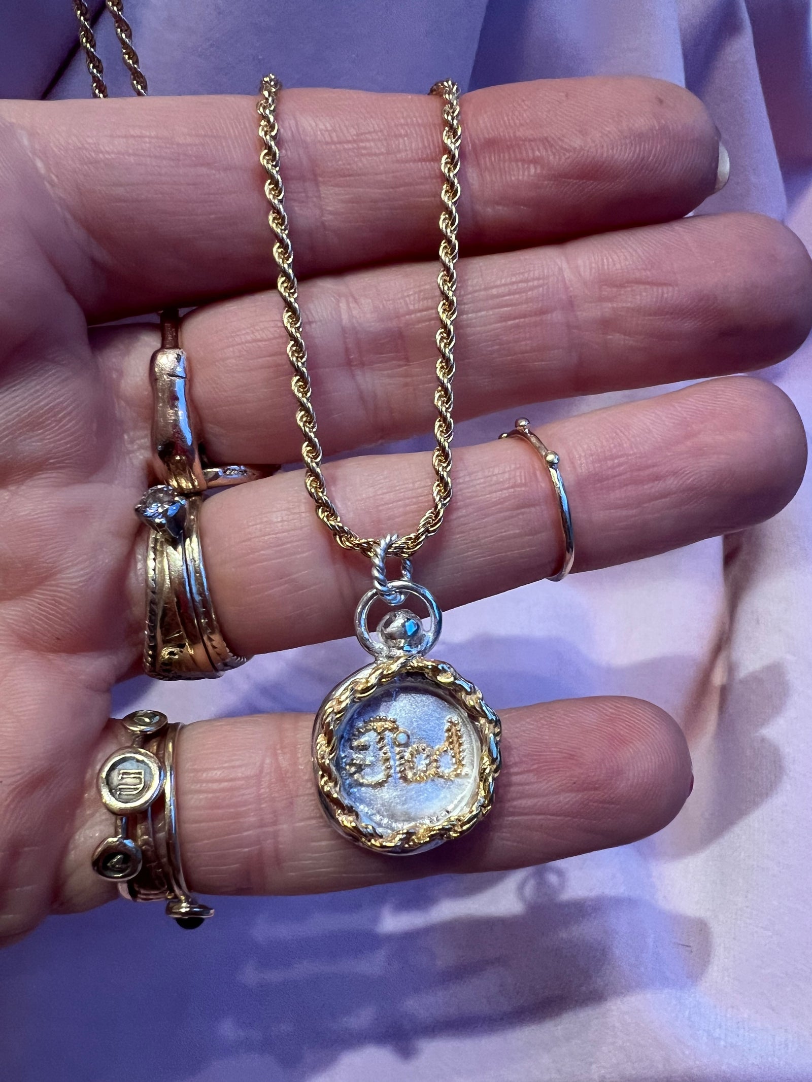 A hand adorned with gold rings holds Jessica de Lotz Jewellery's Tied Charm—a gold chain necklace with a round, wax seal-style pendant engraved with "TIED" on white—resting against pale pink fabric.