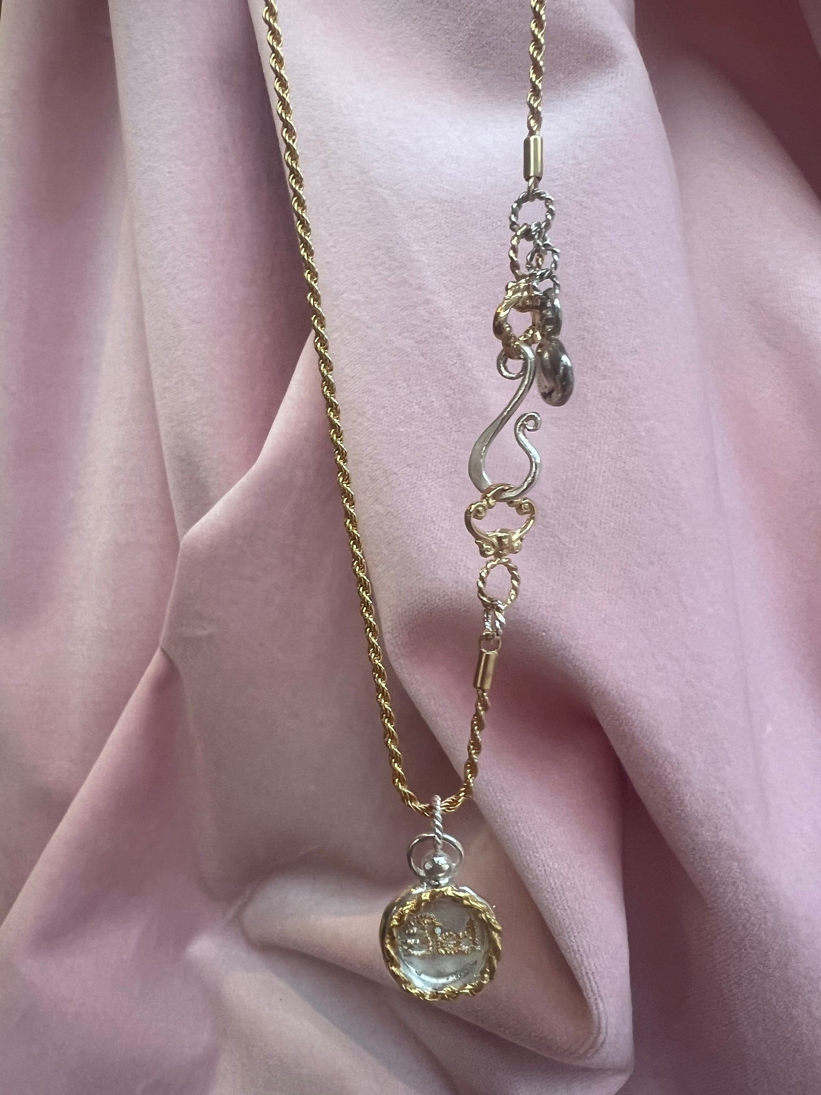 The Tied Charm necklace by Jessica de Lotz Jewellery features a gold rope chain with a round, gold-rimmed "best friend" pendant and decorative silver and gold clasps, displayed on soft pink fabric.