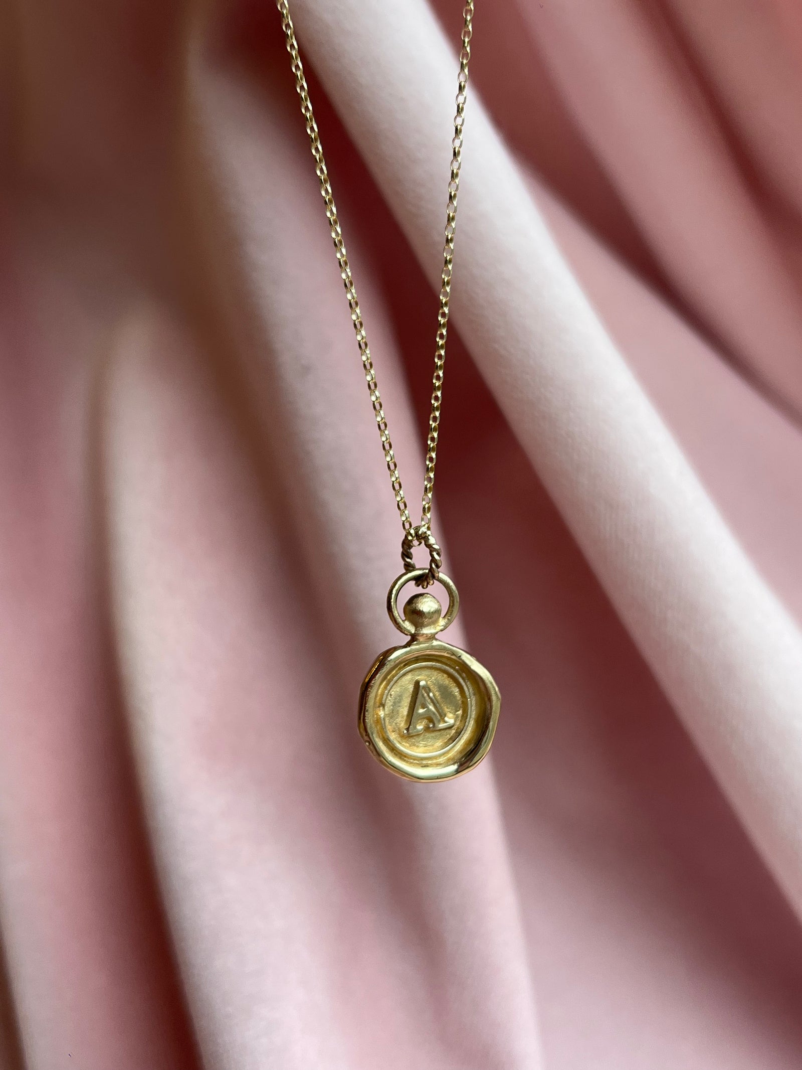 The Mini Personalised Wax Seal Charm by JdL Jewellery is a delicate gold necklace featuring an engraved letter A initial pendant with a vintage-inspired wax seal design, shown here against soft pink velvet fabric.