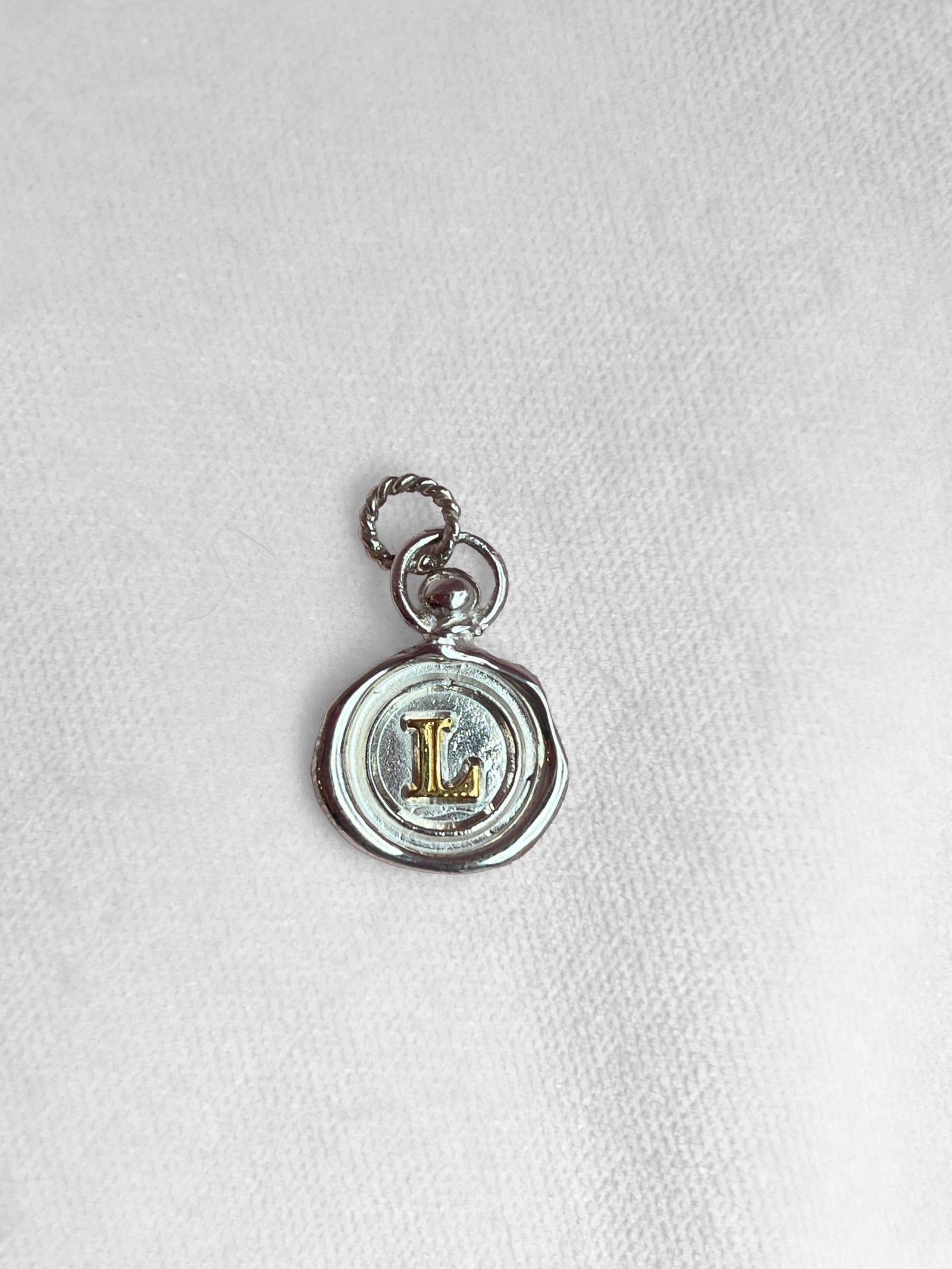 The Mini Personalised Wax Seal Charm by JdL Jewellery features a silver pendant with a twisted loop, textured edges resembling melted wax, and a raised gold “L” at the center, casting a delicate shadow on fabric.