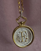 The Large Double Sealed Wax Seal Necklace by Jessica de Lotz Jewellery features a round, gold-plated silver medallion with an ornate letter E and textured silver-toned background, suspended from a delicate gold chain.