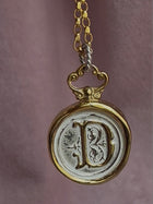 A gold chain from Jessica de Lotz Jewellery features the Large Double Sealed Wax Seal Necklace—a round pendant with a raised, ornate letter D on a silver-toned face framed by gold, set against soft mauve fabric for a vintage touch.