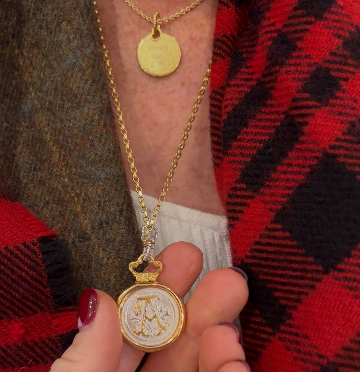 A person wears layered necklaces: one with a small gold disc and another—the Large Double Sealed Wax Seal Necklace by Jessica de Lotz Jewellery. They hold the larger pendant, showing manicured nails, a red and black plaid scarf, and a brown coat.