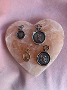 Four St Christopher Charms by Jessica de Lotz Jewellery, in silver and gold, are displayed on a heart-shaped pink and cream marbled stone tray, with a soft pink fabric background highlighting their embossed vintage designs.
