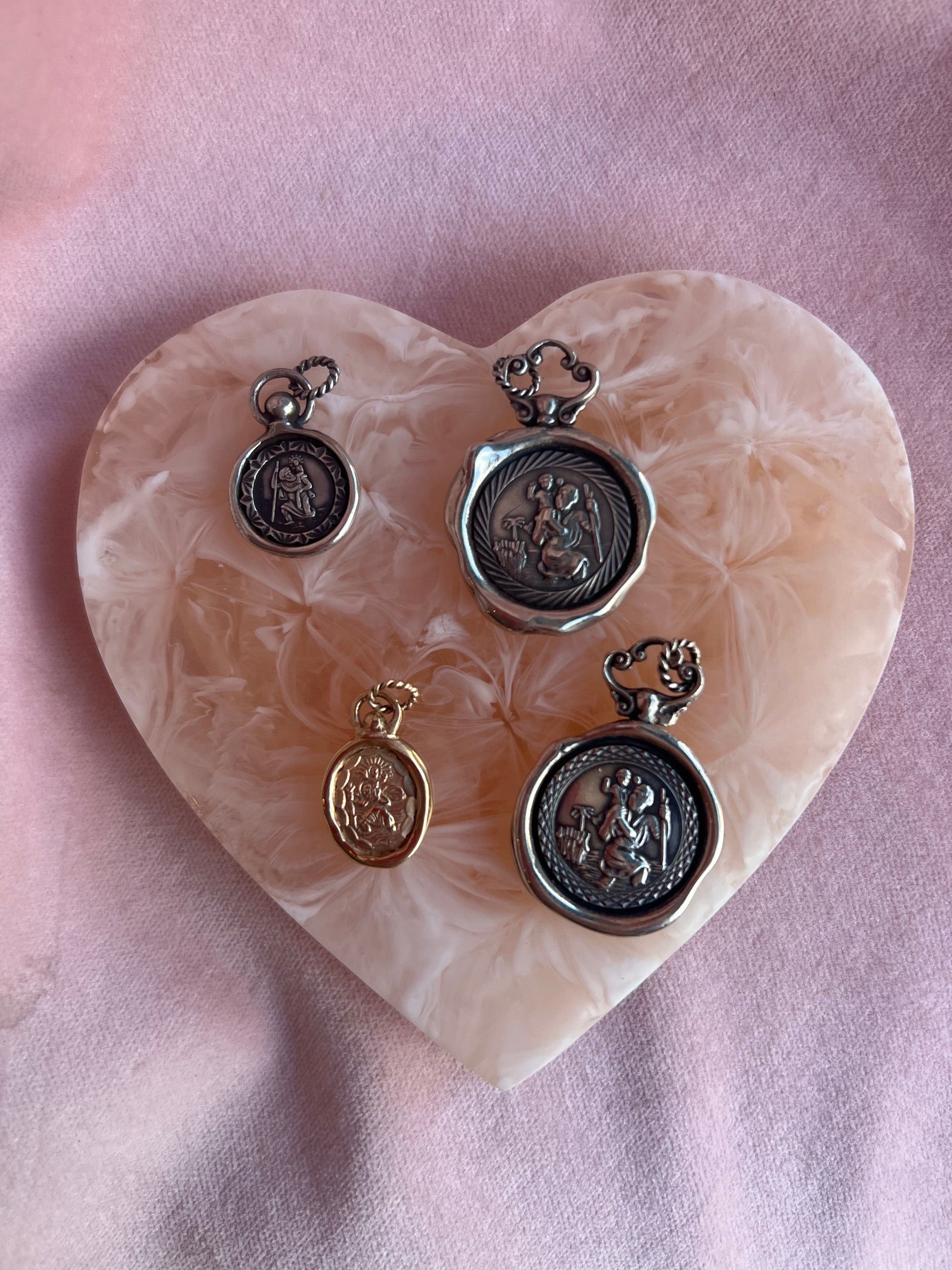 Four St Christopher Charms by Jessica de Lotz Jewellery, in silver and gold, are displayed on a heart-shaped pink and cream marbled stone tray, with a soft pink fabric background highlighting their embossed vintage designs.