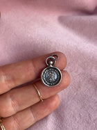 A hand with gold rings holds a small, round St Christopher Charm from Jessica de Lotz Jewellery. The silver pendant features an engraved central figure, a decorative border, and rests on soft pink fabric.