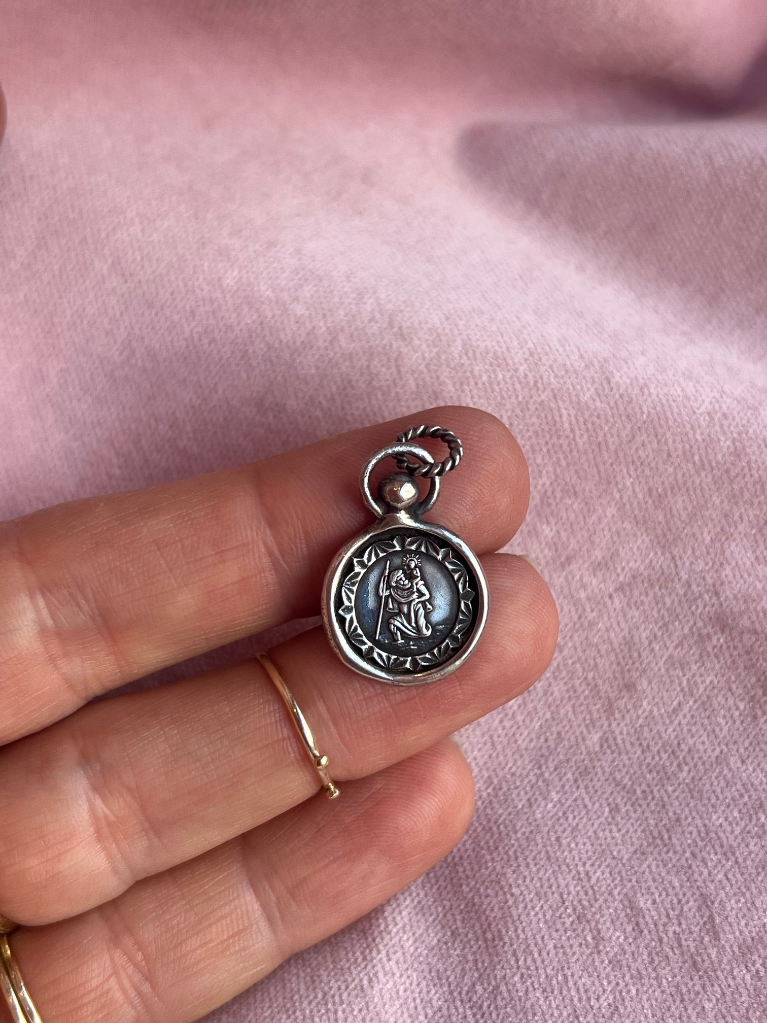 A hand with gold rings holds a small, round St Christopher Charm from Jessica de Lotz Jewellery. The silver pendant features an engraved central figure, a decorative border, and rests on soft pink fabric.