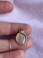 A hand with a gold ring holds an oval St Christopher Charm by Jessica de Lotz Jewellery, featuring a raised face and sunburst design, on light pink fabric. The pendant has a twisted loop at the top for attaching to a chain.